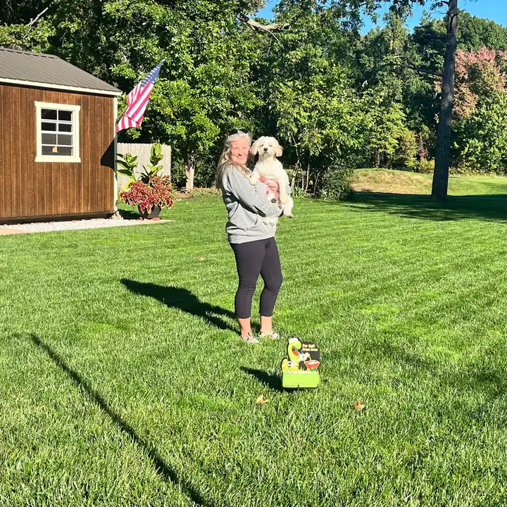 A homeowner holding her dog on a thick, evenly striped lawn beside a shed and flagpole