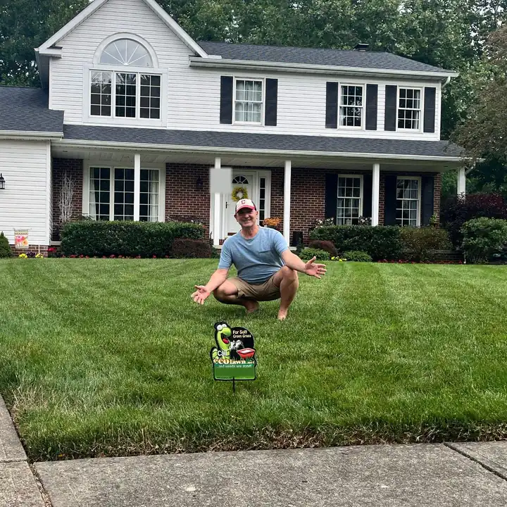 A homeowner crouching with open arms on a thick, freshly striped lawn in front of his home