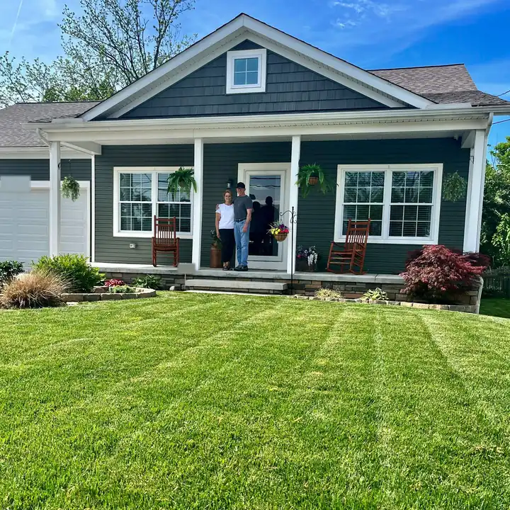 A couple on the front porch of a gray craftsman home with a thick, striped front lawn
